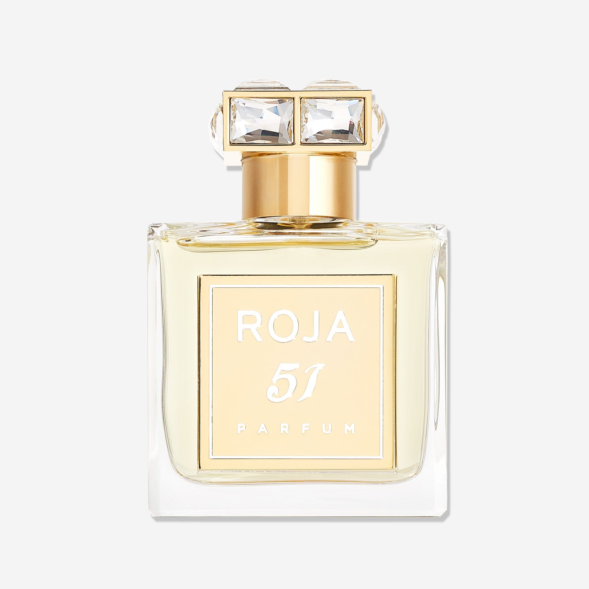 A clear glass bottle of Roja Parfums 51 Pour Femme features a gold label and cap adorned with two large square jewels. The pale yellow luxury fragrance pops against a white background, capturing the elegance of Mayfair London.
