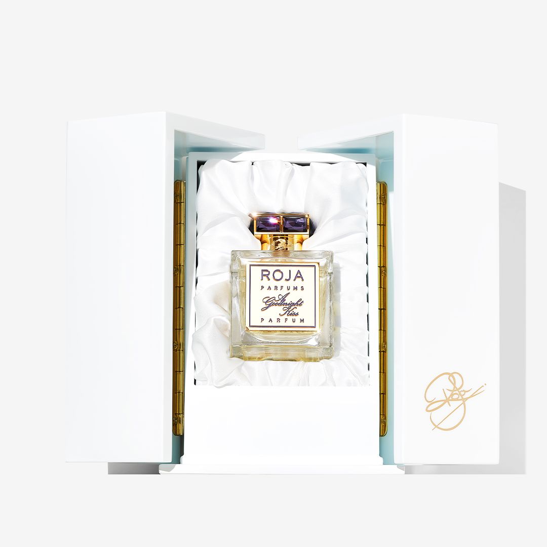 A bottle of Roja Parfums A Goodnight Kiss, a luxury Parfum, is displayed upright in a white, satin-lined, hinged presentation box with gold accents and a signature on the inside of the door.