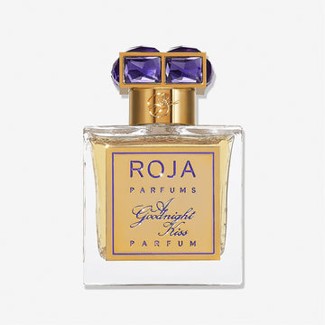 A clear glass bottle of Roja Parfums A Goodnight Kiss, featuring a gold label and an elegant gold cap set with two large purple gemstones.