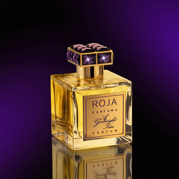 A luxurious gold and glass perfume bottle labeled ROJA Parfums A Goodnight Kiss Parfum stands on a reflective surface with a deep purple gradient background. The cap is gold with purple geometric accents.