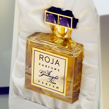 A bottle of Roja Parfums Goodnight Kiss perfume with a gold cap featuring two purple gemstones, nestled in a white satin-lined box.