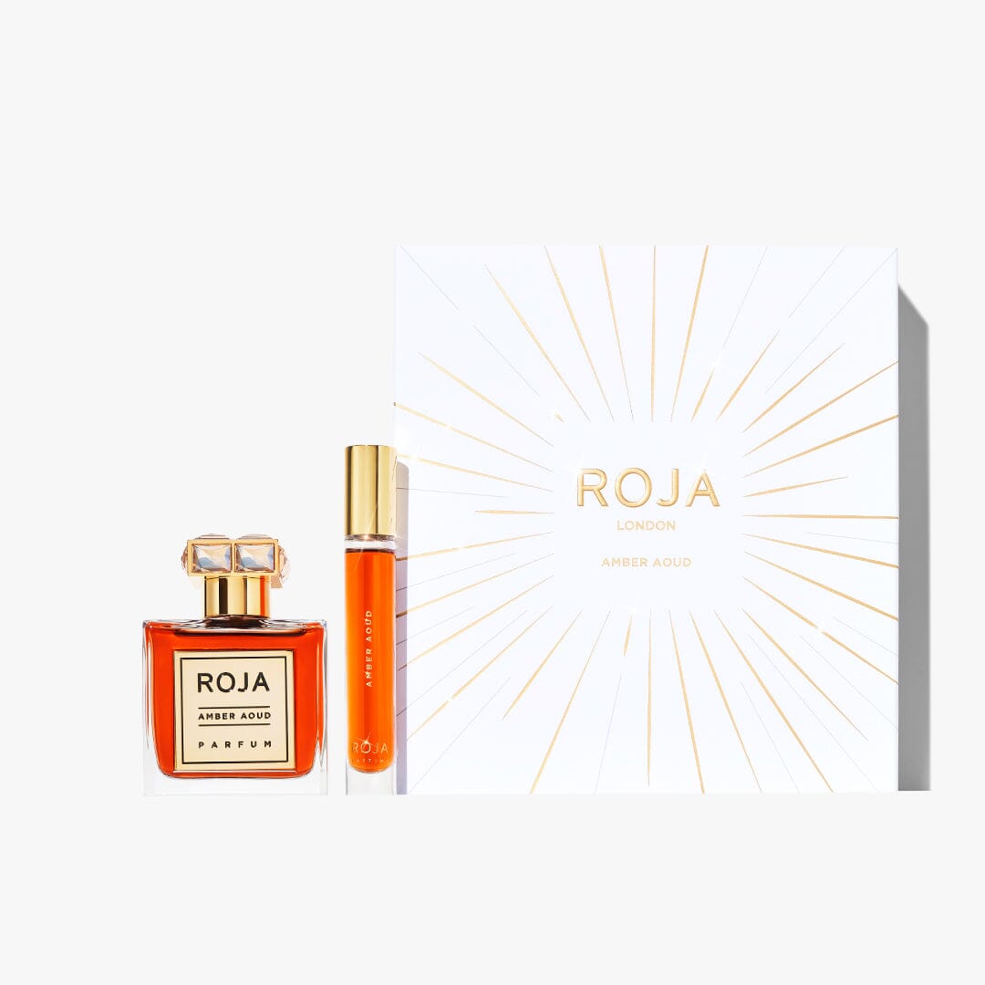 The Roja Parfums Amber Aoud Gift Set includes a square glass bottle, a cylindrical travel spray, and a white box with gold rays and lettering. This elegant oud gift set is beautifully displayed on a light background.