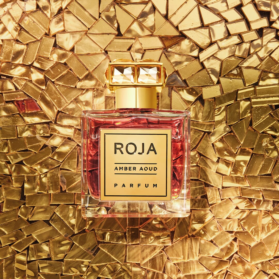 A bottle of Roja Parfums Amber Aoud parfum, featuring a gold cap and label, sits atop a shimmering mosaic-like gold background. The amber-hued fragrance with rich oud harmonizes beautifully with the opulent setting.
