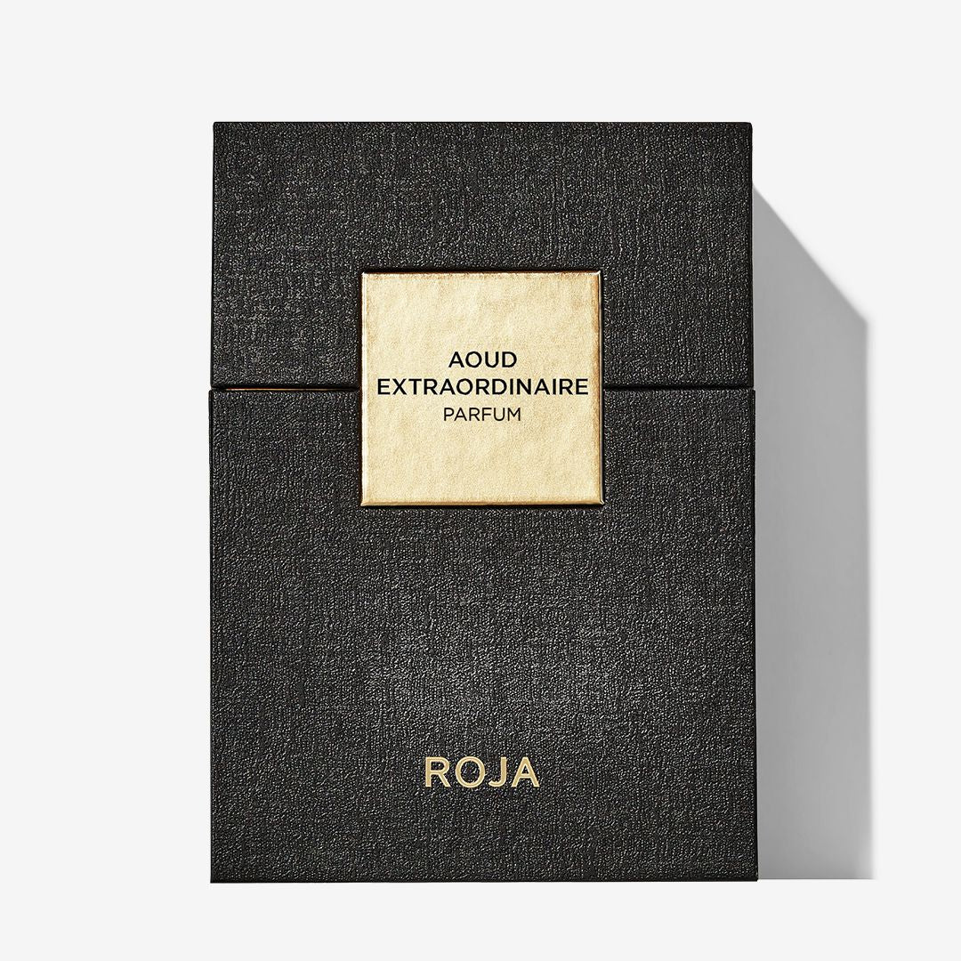 A black textured box with a gold square label reading Aoud Extraordinaire Parfum and Roja Parfums in gold at the bottom sits on a white background, casting a shadow to the right.