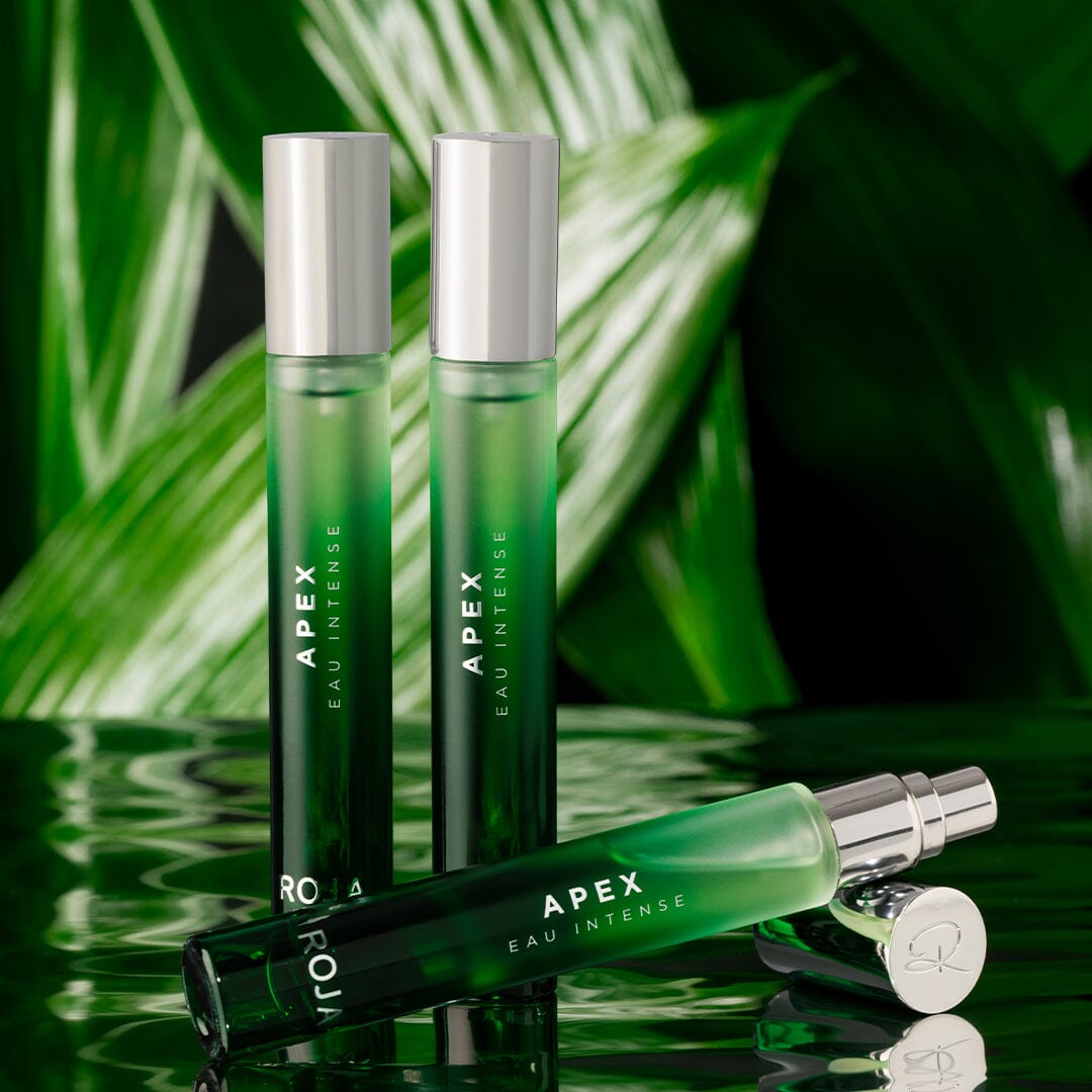 Three green glass bottles of Roja Parfums Apex Eau Intense stand on a glossy, reflective surface amid lush green leaves, reflecting the fragrance’s fresh atmosphere and signature Chypré accord.