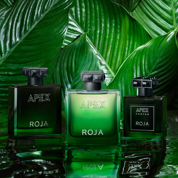 Three bottles of Roja Apex perfume in green and black sit side by side in front of large, glossy green tropical leaves, creating a lush and luxurious setting.