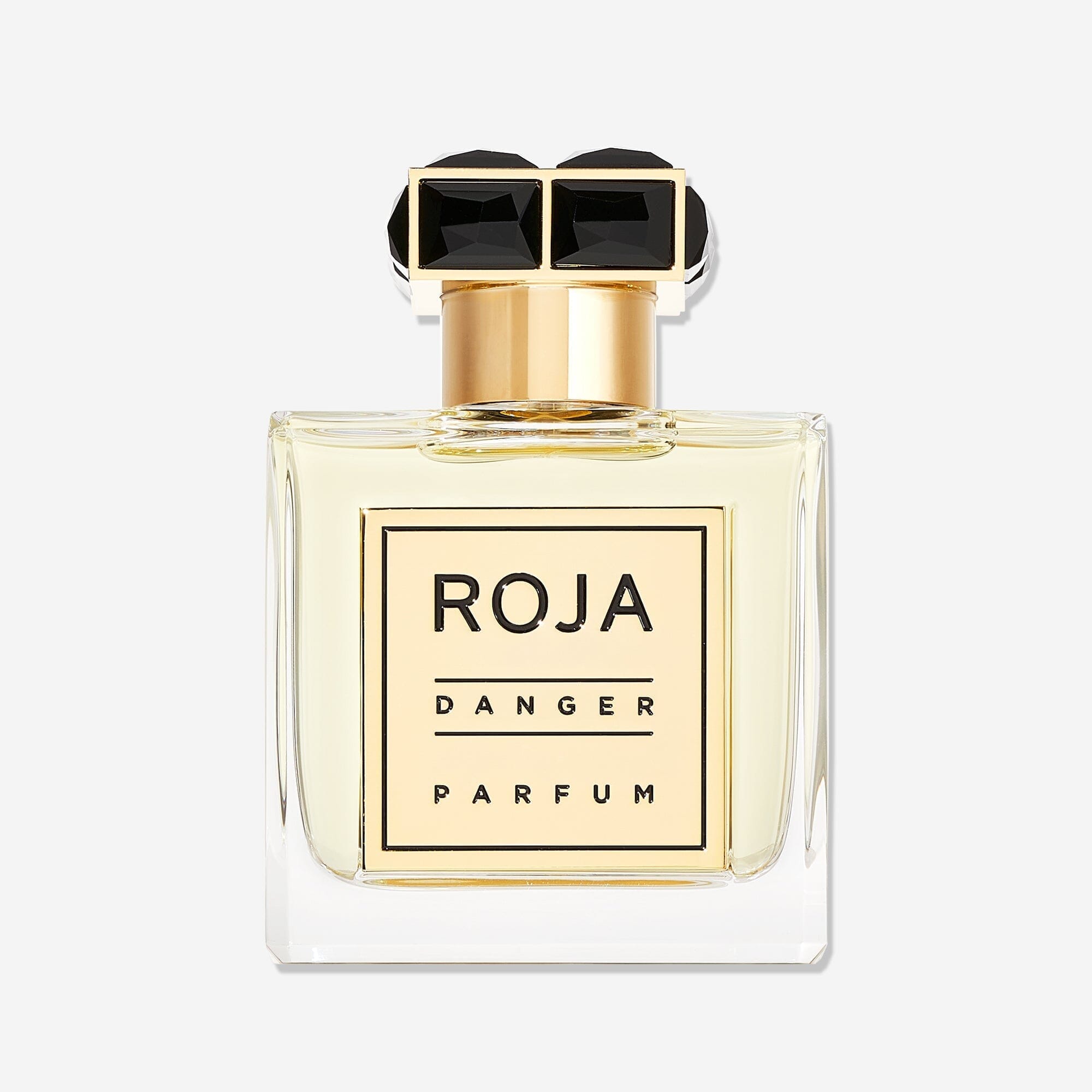 A clear glass bottle of Roja Parfums Danger Pour Homme, with a gold label and black text, topped by a gold cap adorned with two black gem-like shapes. The light yellow liquid inside is a seductive men’s fragrance.