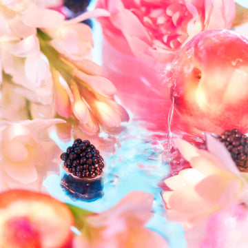 A close-up of blackberries, peaches, and pink and white flowers on a reflective surface evokes the dreamy, Elysium-inspired aesthetic of Roja Parfums Elysium Pour Femme.