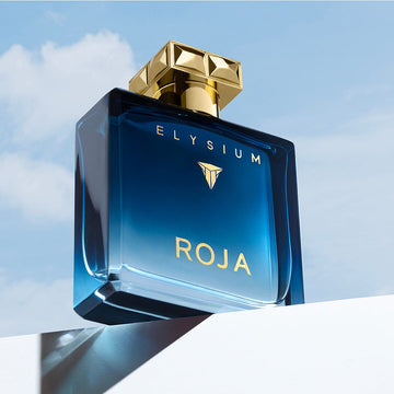 A blue glass bottle of Roja Parfums’ Elysium Coffret, featuring a gold cap and lettering, stands against a sky with clouds on a white surface—an elegant display for its bold masculine scent.