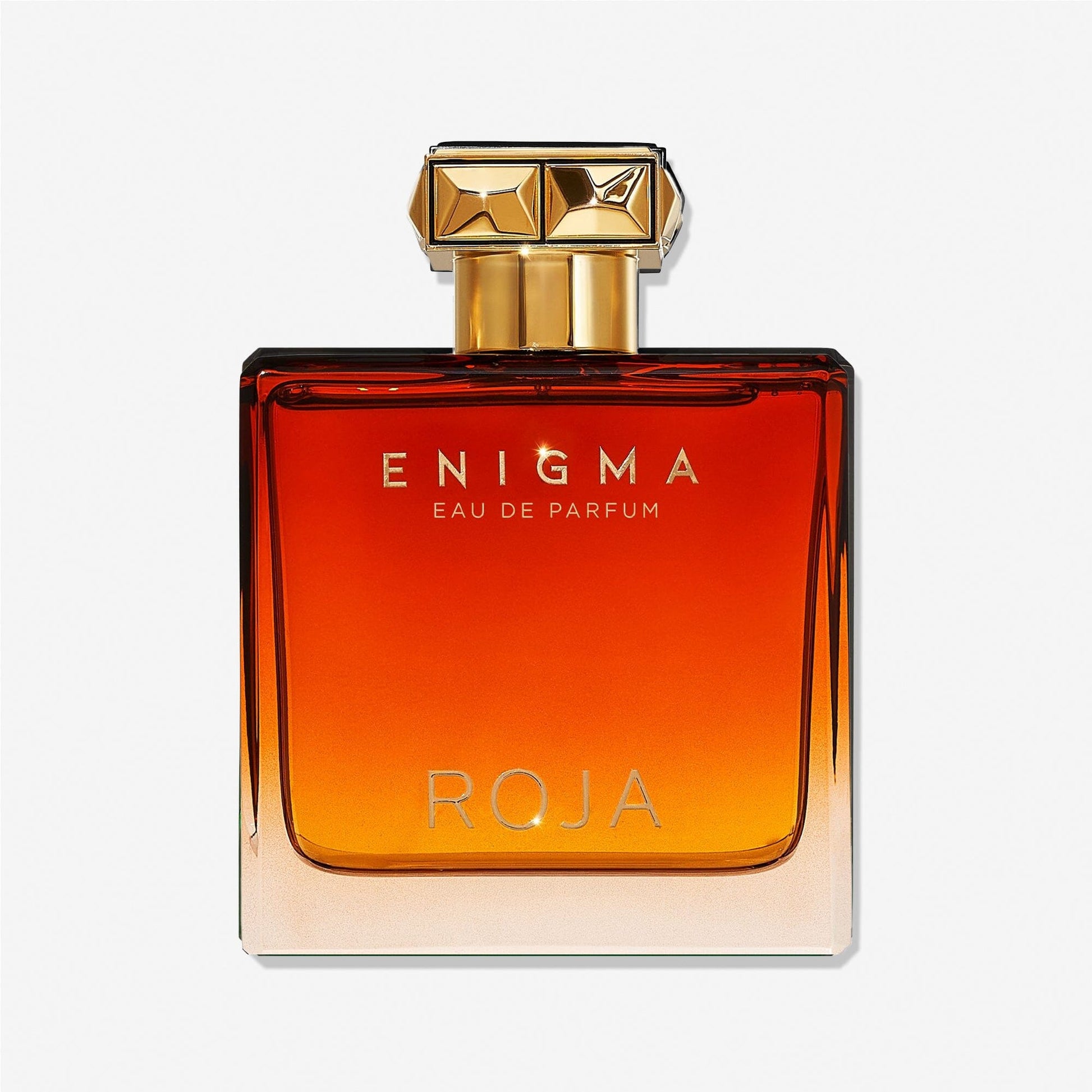 A rectangular glass bottle featuring a gradient amber-to-clear color, gold cap with geometric design, and gold â€œEnigma Pour Homme Roja Parfumsâ€ text on the front, evoking rich tobacco and smooth vanilla notes.