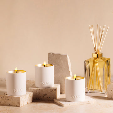 Three lit white candles with gold interiors, labeled ROJA, are placed on stone blocks beside a glass diffuser with wooden reeds, all set against a neutral beige background with stone decor elements.