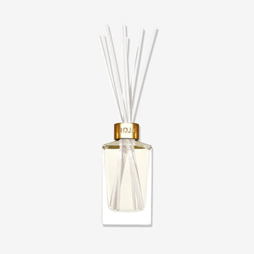 A clear glass reed diffuser labeled "London" by Roja Parfums, featuring a gold cap, light-hued liquid, and white reeds, exudes elegance against a white background.