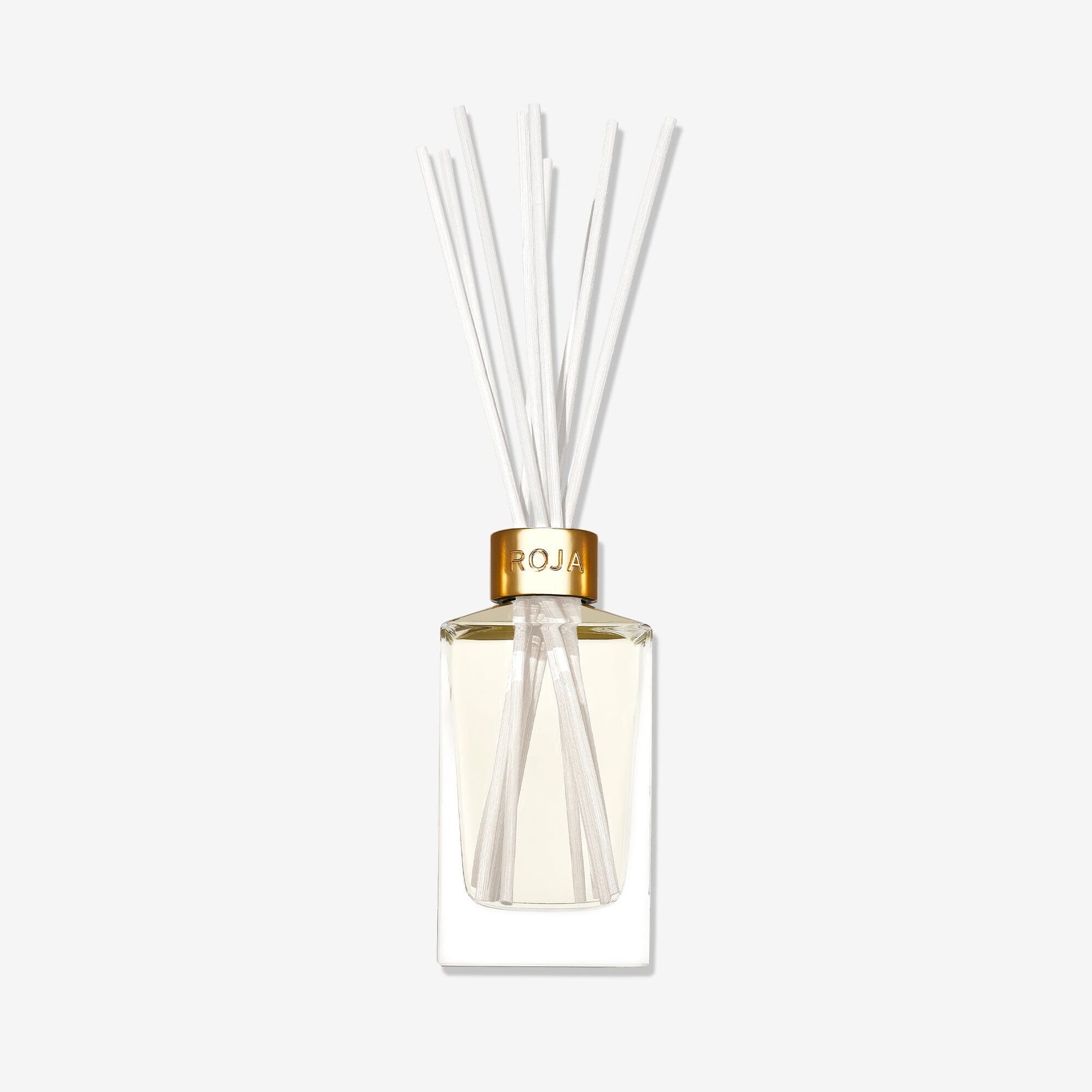 A clear glass reed diffuser labeled "London" by Roja Parfums, featuring a gold cap, light-hued liquid, and white reeds, exudes elegance against a white background.