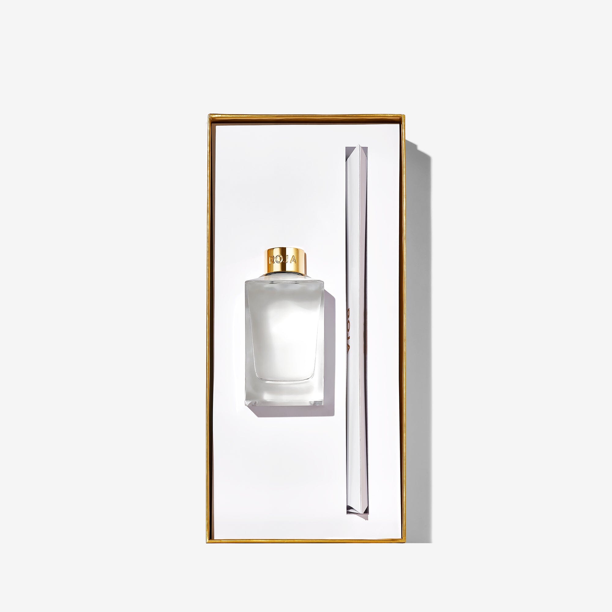 A clear Roja Parfums "London" perfume bottle with a gold cap and matching gold-trimmed box stands beside a slim white item, embodying London elegance against a plain white background.