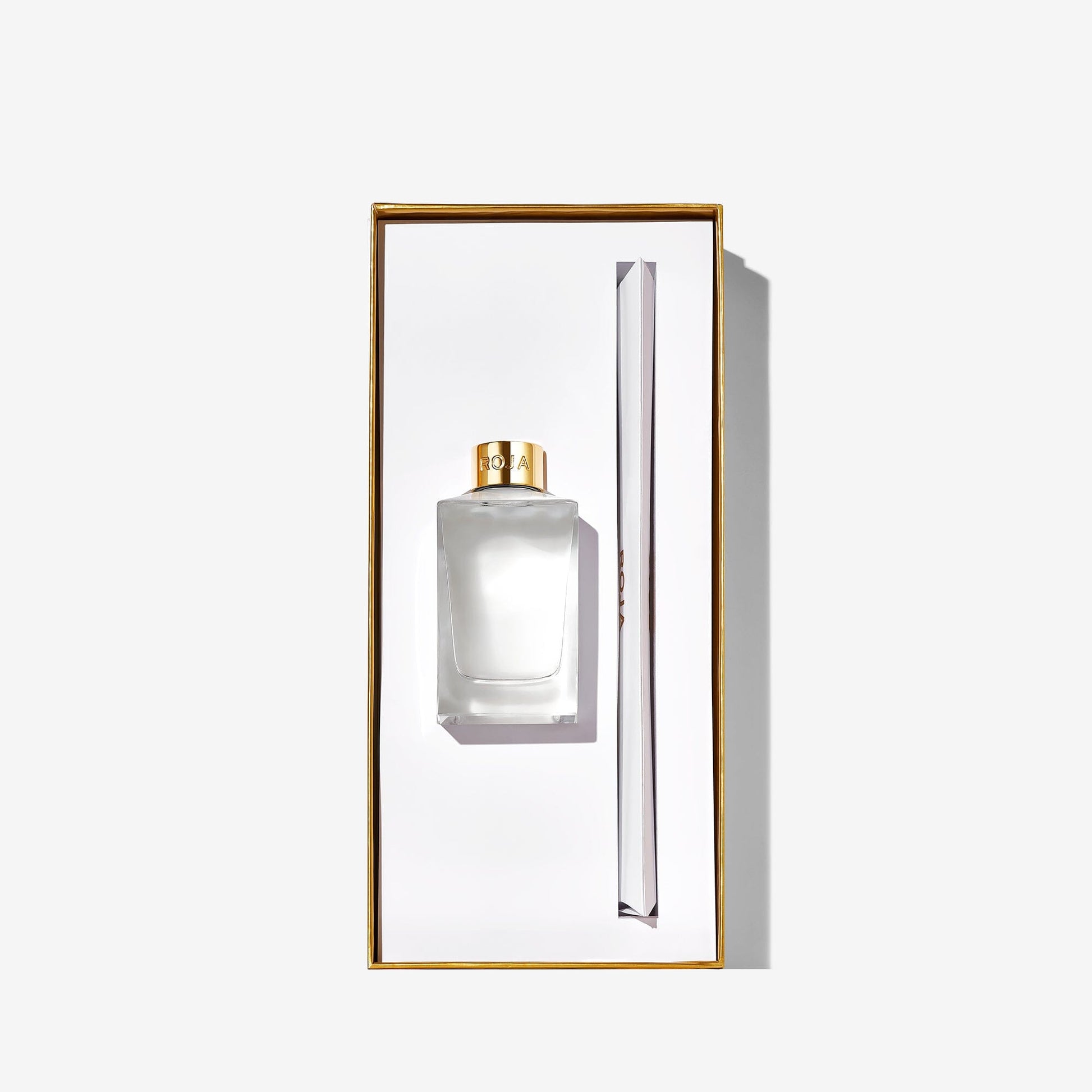 A clear Roja Parfums "London" perfume bottle with a gold cap and matching gold-trimmed box stands beside a slim white item, embodying London elegance against a plain white background.