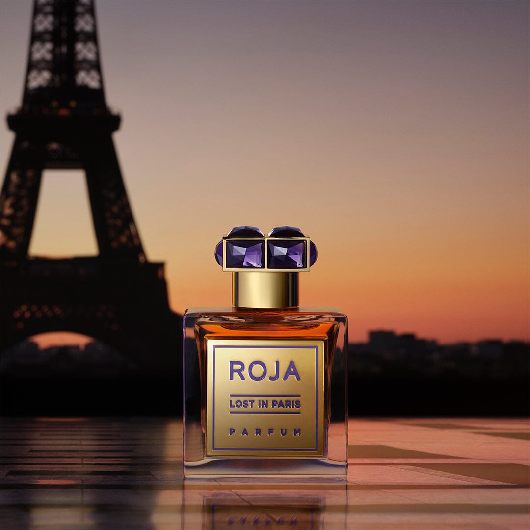 A bottle of Roja Lost in Paris parfum sits in the foreground with the Eiffel Tower softly blurred in the background at sunset. The bottle features a gold label and a gem-like cap.