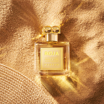 A square glass perfume bottle labeled ROJA ISOLA SOL PARFUM sits on golden sand, illuminated by warm sunlight, with textured fabric visible in the lower left corner.