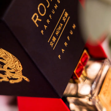 A close-up of a luxury Roja Parfums NÜWA perfume box in black and gold with a dragon design, partially open to reveal the glass perfume bottle inside with a gold label and red accents.