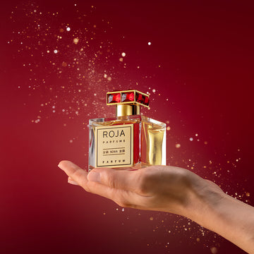 A hand holds a luxurious Roja Parfums perfume bottle with a gold label and red gemstone cap, set against a rich red background with golden sparkles.