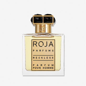 A clear glass bottle of Roja Parfums Reckless Pour Homme features a gold and black cap and holds light yellow liquid. The label shows the brand and product name in bold black lettersâ€”a masculine scent with spicy chyprÃ© notes.
