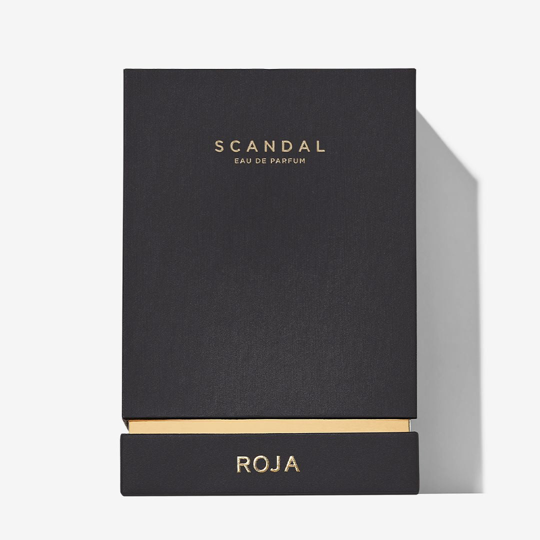 A sleek black rectangular box with gold text displays SCANDAL POUR HOMME at the top and ROJA PARFUMS at the bottom on a pulled-out drawer, suggesting a refined menâ€™s cologne inspired by classic barbershop traditions.