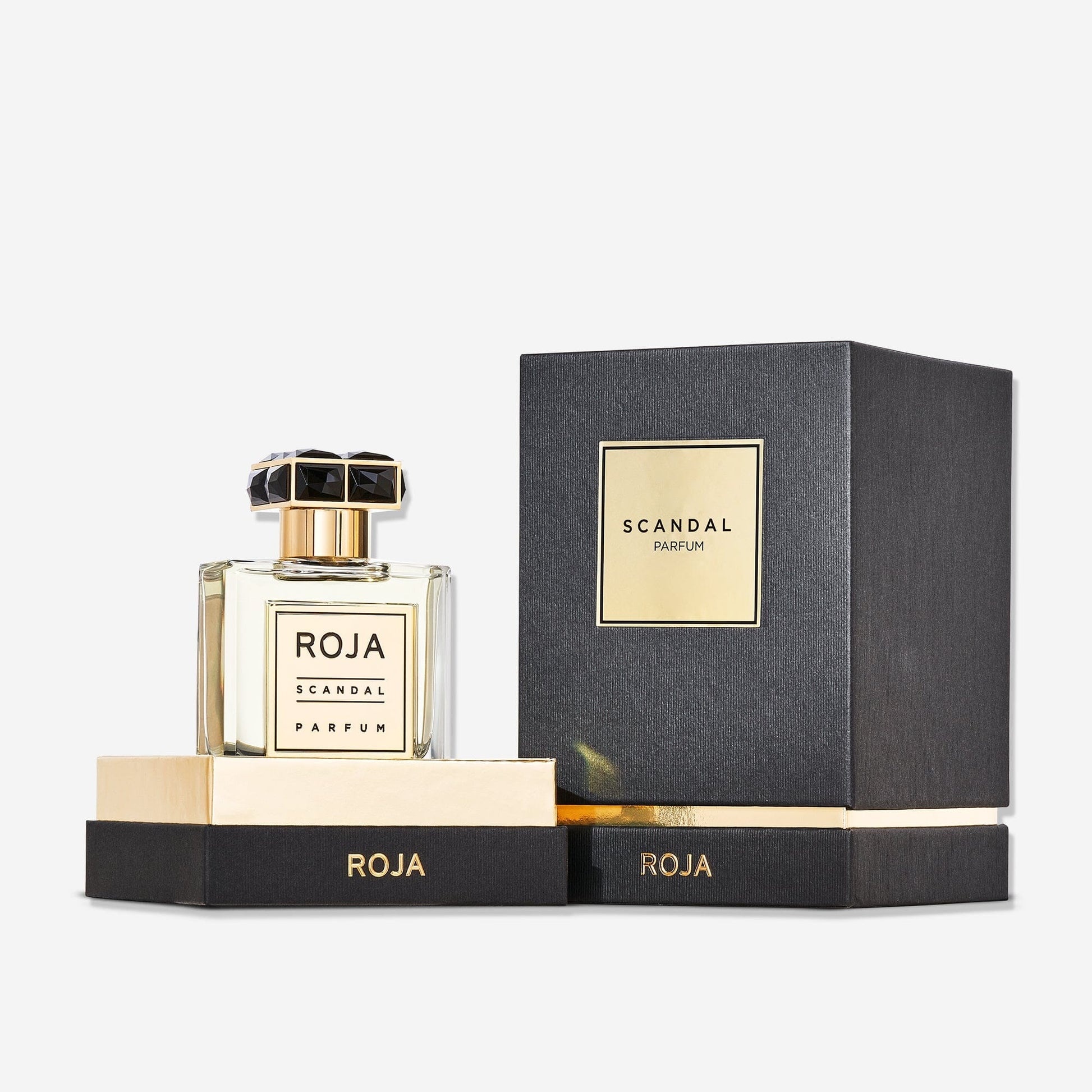 A bottle of Scandal Pour Homme by Roja Parfums rests on a gold and black base beside its matching box, both adorned with minimalist, elegant labelsâ€”a sophisticated FougÃ¨re scent for todayâ€™s gentleman.