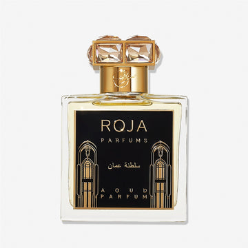 A clear perfume bottle labeled Roja Parfums Sultanate of Oman features gold accents, a black and gold label with Arabic designs, and a crystal-embellished gold capâ€”evoking luxurious Aoud and smoky frankincense.