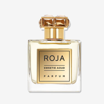 A clear glass bottle of Roja Parfums Sweetie Aoud features a gold label and gold cap with two large, square, amber gemstones. The bottle holds a light amber liquid, highlighting this luxurious Sweet Oud perfume.