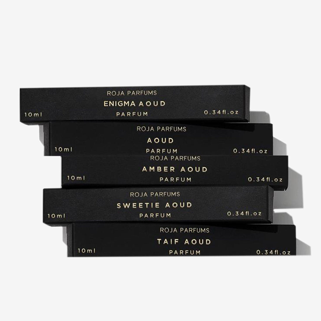 Five sleek black perfume boxes with gold lettering are stacked askew. The Aoud Travel Collection by Roja Parfums includes 10ml scents: Enigma Aoud, Aoud, Amber Aoud, Sweetie Aoud, and Taif Aoud.
