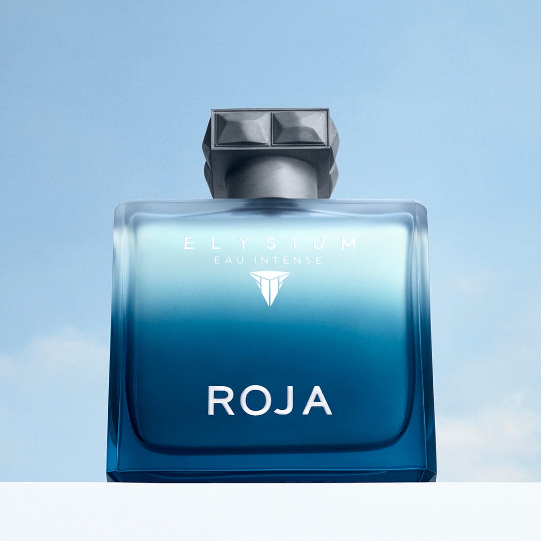 A blue gradient bottle from The Elysium Trio by ROJA London stands against a light sky backdrop, featuring bold white branding and a geometric silver cap—a striking addition to any fragrance collection.