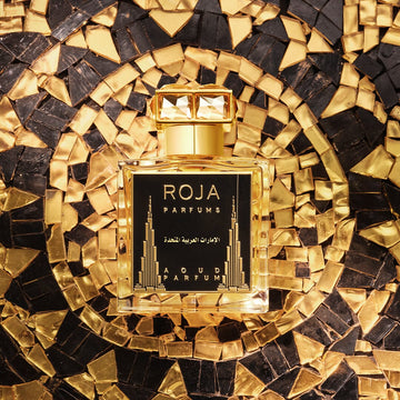 A gold and black bottle of Roja Parfums Aoud Parfum is placed on a shimmering, mosaic gold and black patterned surface. The bottle features Arabic writing and stylized skyscraper designs on its label.